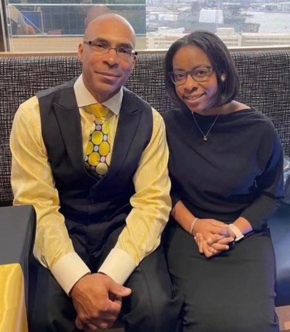 A man in a yellow shirt and patterned tie with a black vest sits beside a woman in a black dress. Both are seated on a bench, holding hands, and smiling at the camera. A cityscape is visible behind them.