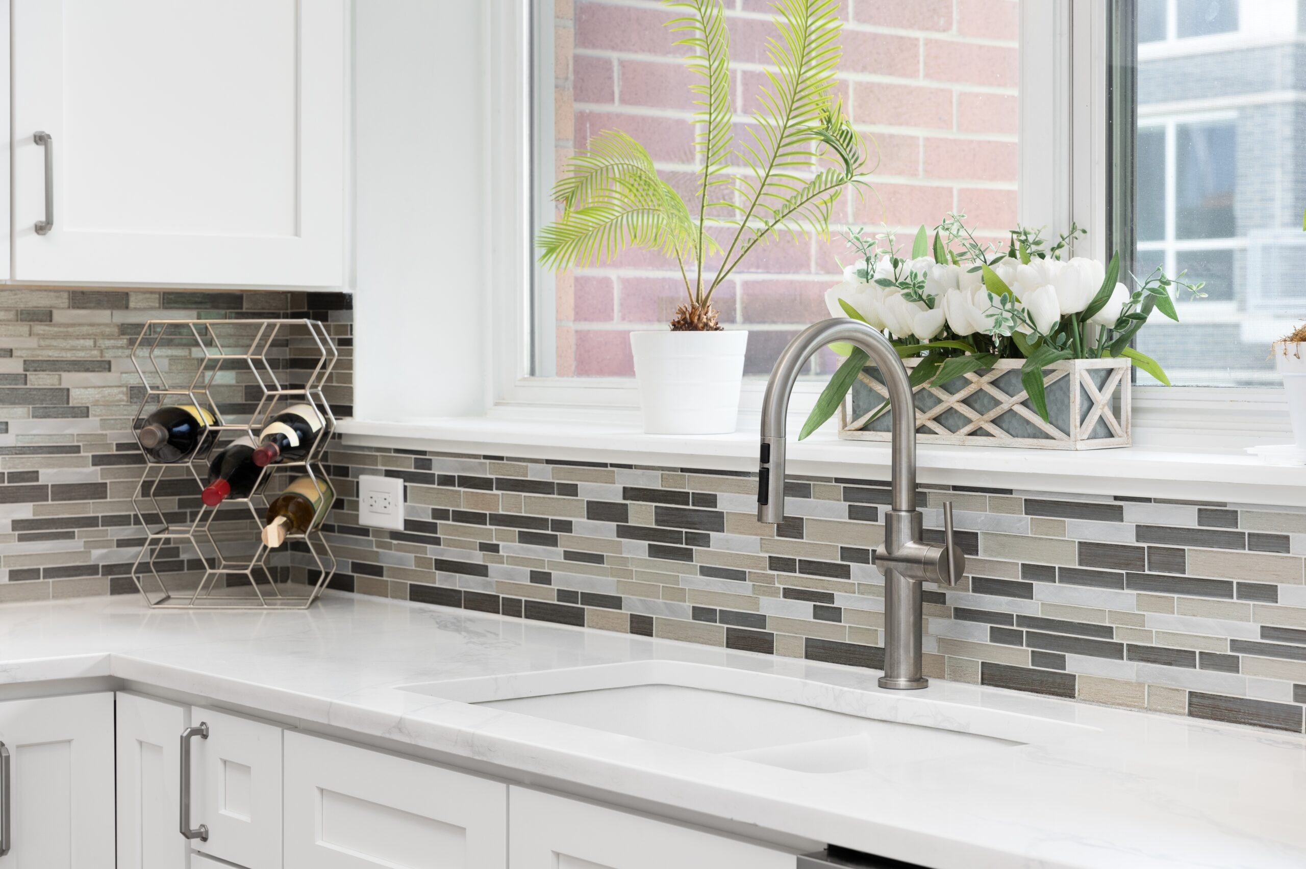 A modern kitchen sink area with a stainless steel faucet, white cabinets, a mosaic tile backsplash, wine rack, and potted plants on a windowsill.